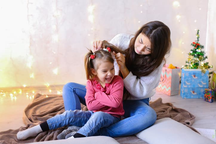 Beautiful girl and elder sister of small female child cares for younger sister and talks, hugs and braids up hair on head, fools around and laughs cute in family way, sitting on floor in bright room against backdrop of glowing garland and small festive tree on eve of new year. Woman with long dark hair of European appearance dressed in white sweater with long sleeves and blue jeans, small female brunette with two tails on each side dressed in raspberry Sweater and blue jeans. Concept of happy and carefree childhood, family values and holidays together, bright and unforgettable New Year's children's emotions, Christmas gifts and Santa Claus, children's entertainment and fun games, decor and bright festive interior.