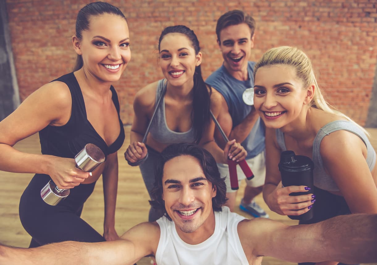 Beautiful sports team are holding sports equipment, looking at camera and smiling while doing selfie in fitness hall