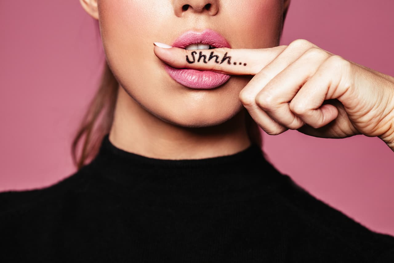 Shh! Women's secrets. Cropped shot of female with finger in mouth. Closeup portrait of young woman is showing a sign of silence with shhh written on the finger.