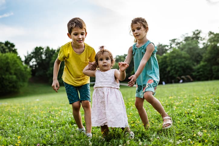 Boy and two girls are in the meadow field. Older siblings are helping baby girl to make first steps.