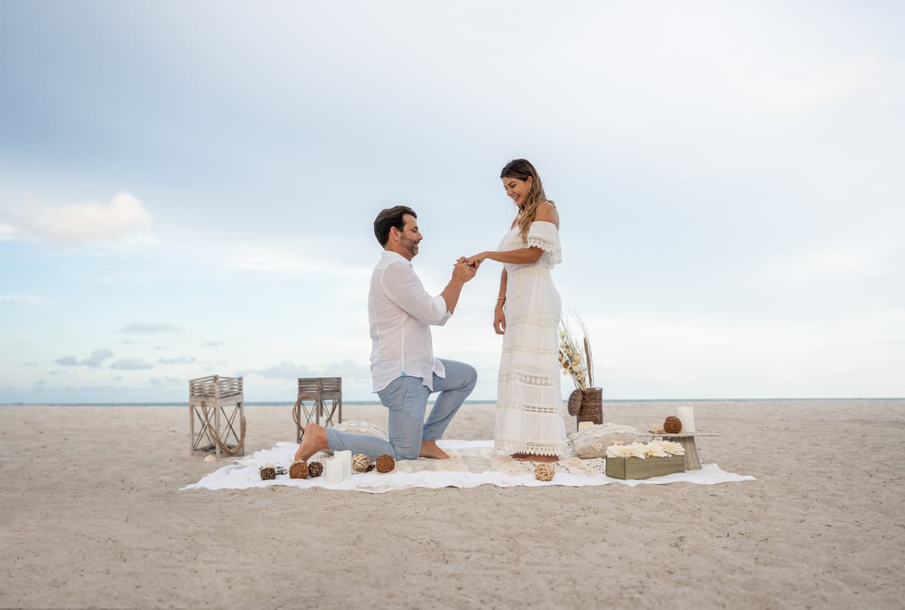 Loving couple getting engaged on a romantic picnic at the beach and him on his knees while he puts the ring on her finger - relationship concepts