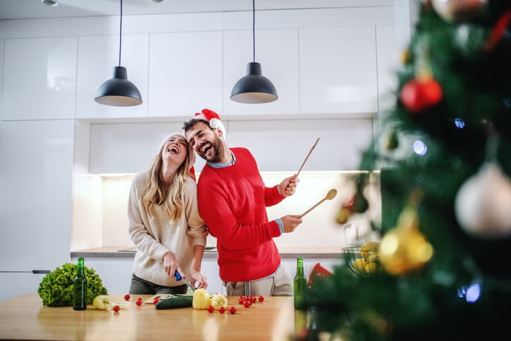 Playful happy caucasian couple with santa hats on heads standing in kitchen and preparing dinner for new year's eve. In foreground is christmas tree.