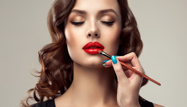 Graceful hands of professional make-up master, with the blue manicure on the nails, is painting in the red color lips of splendid young woman. Creation of perfect make up in progress. Makeup,manicure and cosmetic.