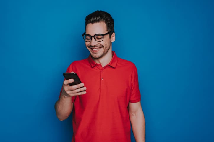 Half length of smiling handsome guy in glasses looking at screen of mobile phone isolated on blue background. He is looking at device and laughing indoors. Human emotions and face expressions concept