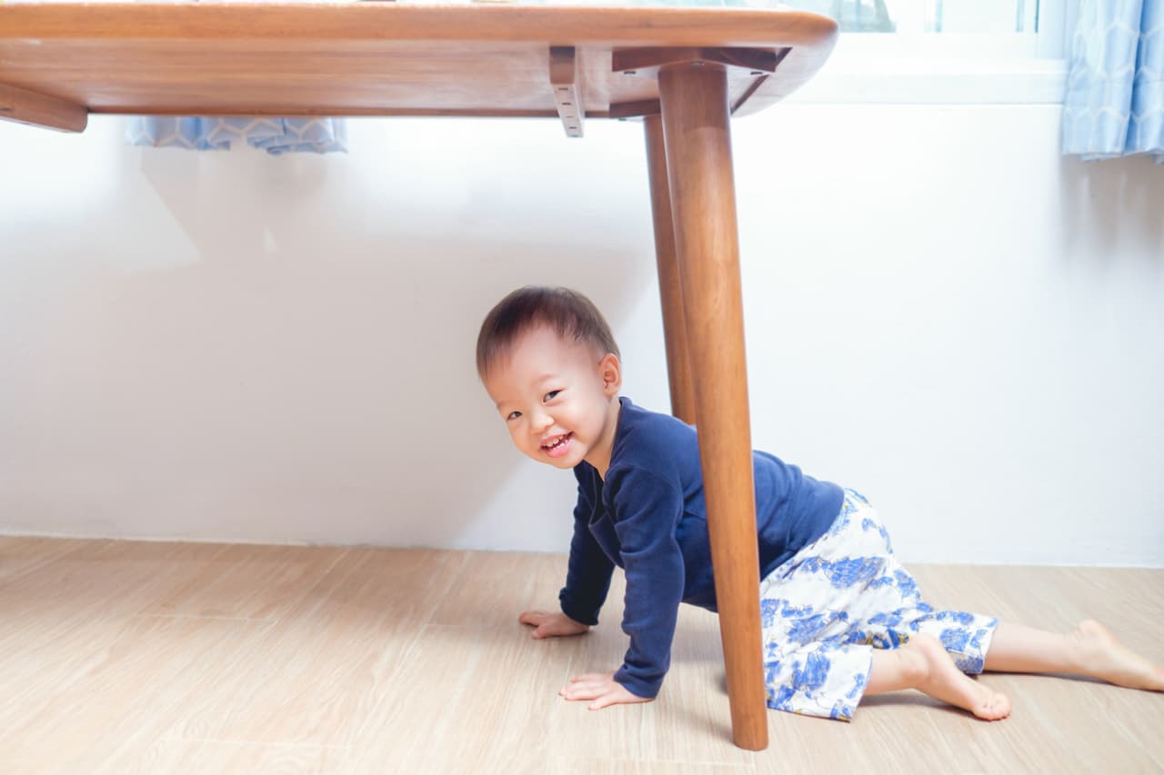 Cute smiling funny little Asian 18 months / 1 year old toddler boy child playing under table at home looking at camera, kid has playful expression on his face, Happy childhood concept