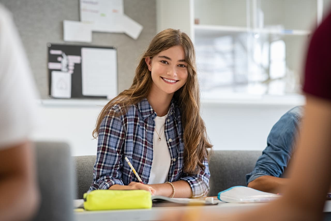 Beautiful university student smiling while studying for exams in classroom. Pretty woman sitting in classroom full of students during class. Portrait of happy young woman writing notes and looking at camera.