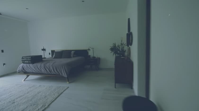 La habitación cuenta con una amplia cama con un buró, una lámpara y una vela, todo en tonalidades oscuras, además de contar con un gran espejo.