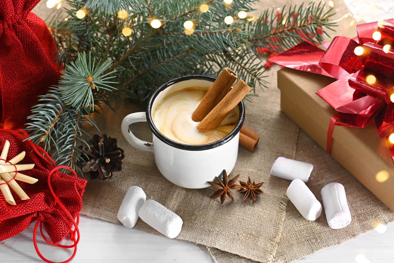 Eggnog with cinnamon, anise and nutmeg in a white mug on a wooden table near the branches of a Christmas tree and decorative toys. Christmas and winter holidays. Greeting card or banner for the site.