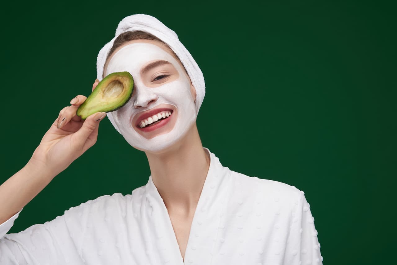 Close up portrait of charming young woman with towel on her head hiding eye behind avocado half and smiling. Isolated on green background