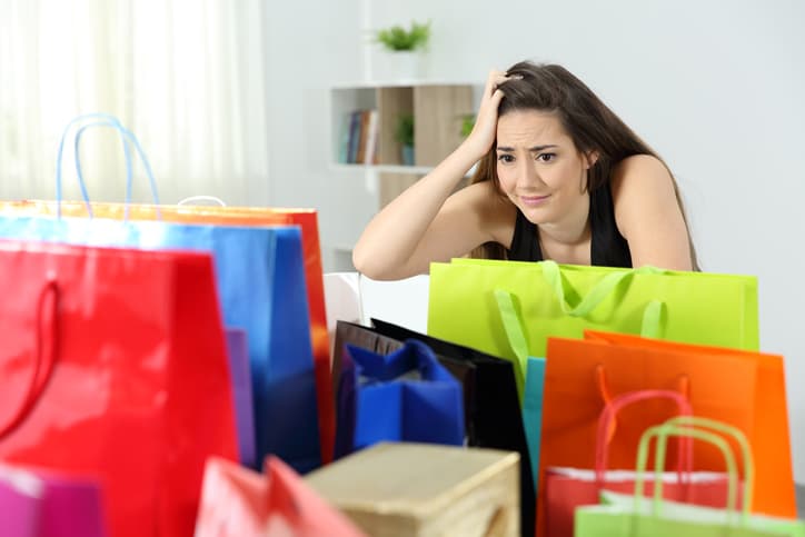 Worried shopaholic woman after multiple purchases in colorful shopping bags at home
