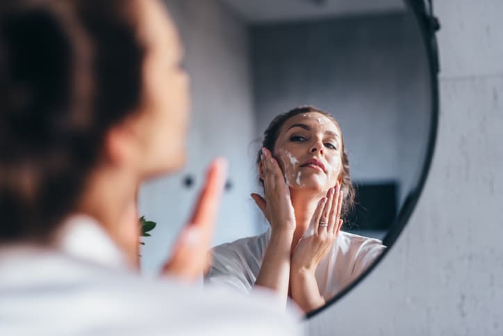 Woman washes her face in front of the mirror, applying foam to her cheeks.