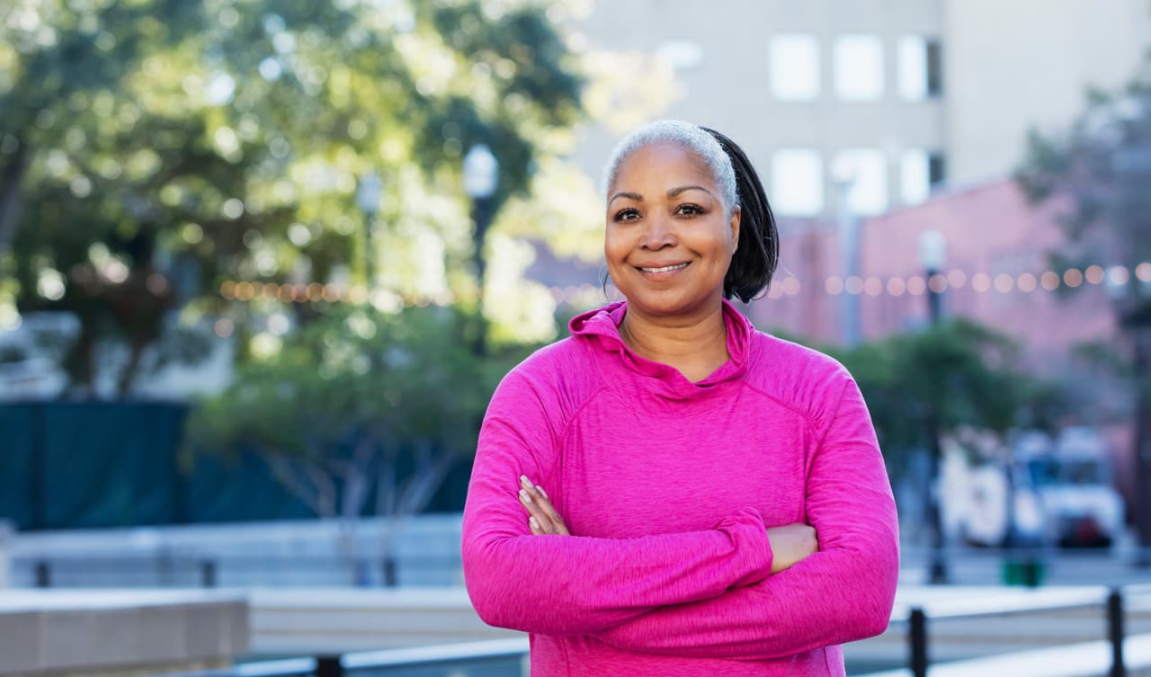 A mature African-American woman in her 50s wearing a pink hooded sweatshirt, smiling as she stands outdoors in the city, a tall building out of focus in the background.