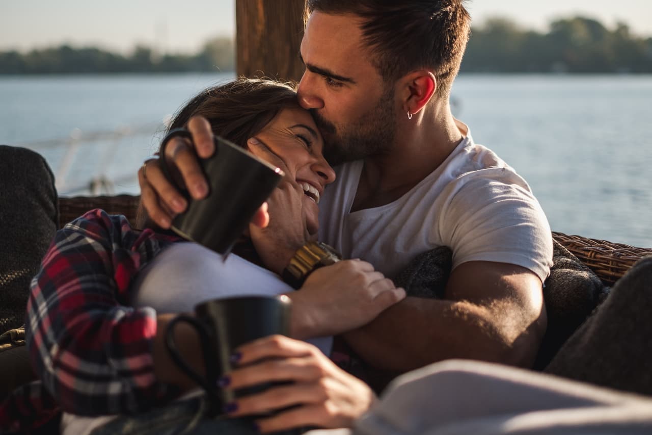 Boyfriend kissing his smiling girlfriend in forehead while she is lying covered with blanket by the river. Beautiful autumn day