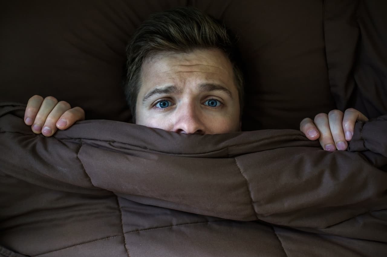 Caucasian young man hiding in bed under the blanket at home. He is stressed and tries run away from problems